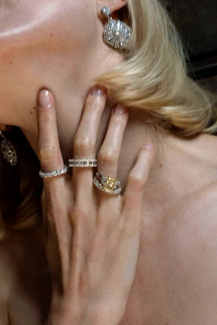A hand at the neck wearing stacked diamond eternity bands and a yellow diamond ring, with diamond drop earrings