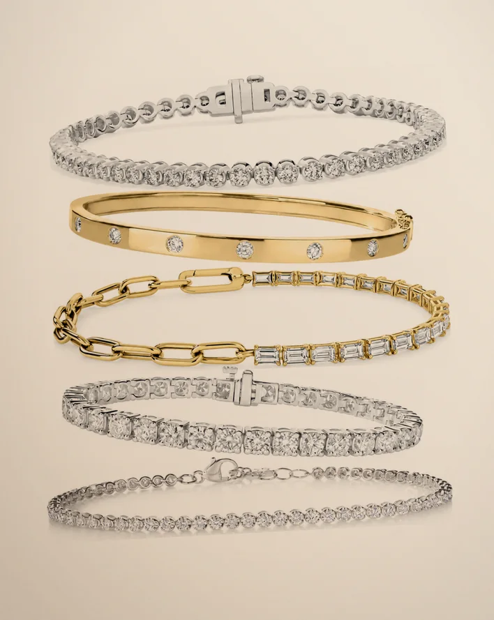 A stack of five diamond bracelets on warm beige paper, from tennis bracelets to a gold inlay bangle and a baguette link chain