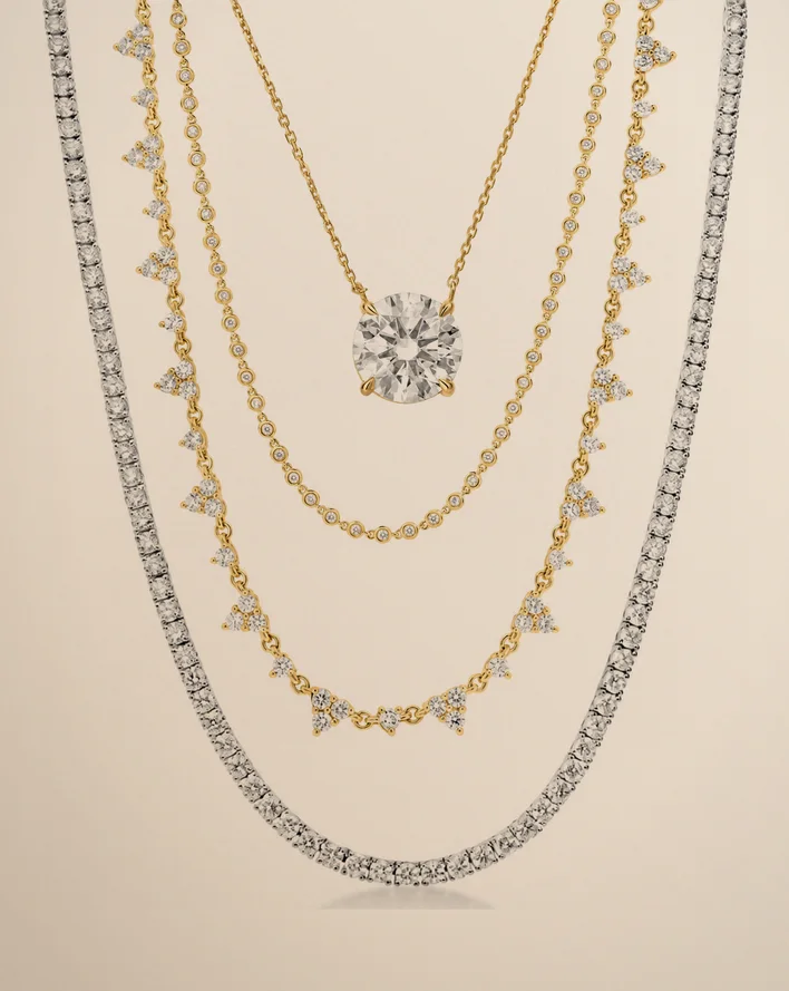 Four diamond necklaces layered on warm beige paper: a tennis necklace, a trio-station necklace, a bezel necklace and a solitaire pendant