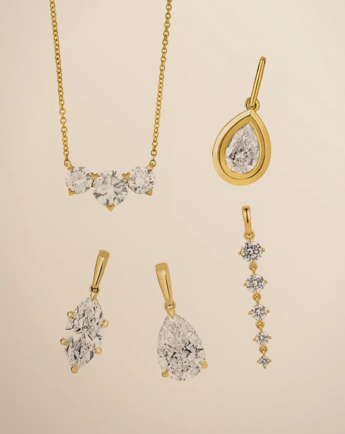 Gold diamond pendants on warm beige paper: a three-stone cluster pendant necklace with a bezel pear, a marquise, a pear and a graduated drop